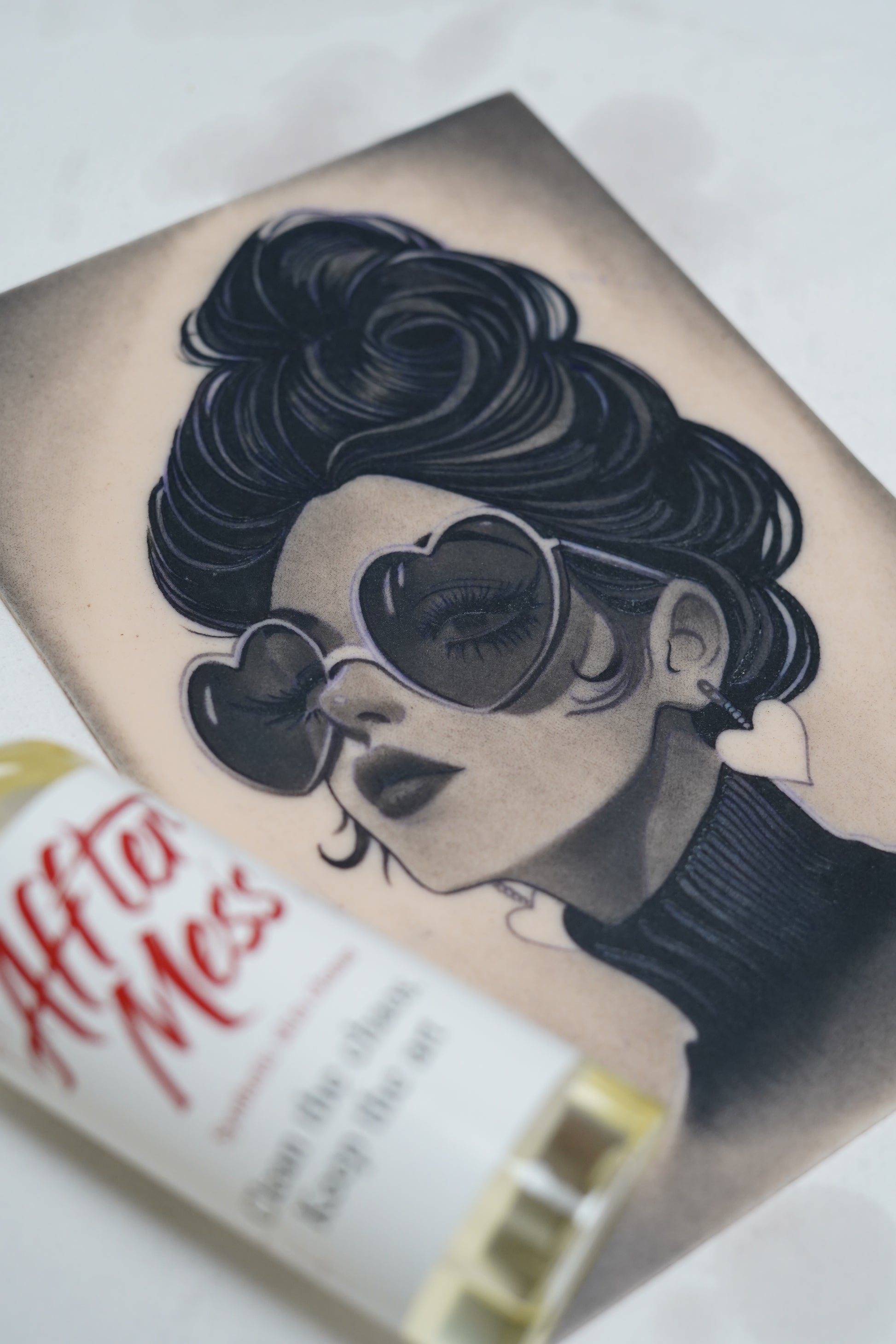 Tattoo of a woman with sunglasses on a person's arm, with a book titled 'After Hours' in the background.